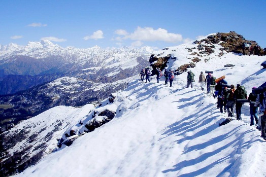 Group of trekkers walking through snowy Himalayan trails on the Kedarkantha Trek with clear blue skies and mountain peaks in the background